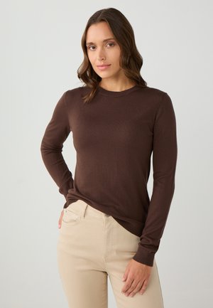 Maglione marrone a maniche lunghe con scollatura rotonda, texture liscia e polsini a coste, indossato sopra pantaloni beige chiaro. Design semplice e aderente.