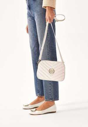 Quilted weiße Handtasche mit einem Logo in Goldtönen, verstellbarem Tragegurt und Reißverschluss; kombiniert mit blauer Jeans und weißen Ballerinas.