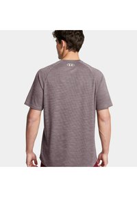 Camiseta deportiva de manga corta, gris, con un patrón texturizado, mangas raglán y un logo en la parte superior de la espalda.