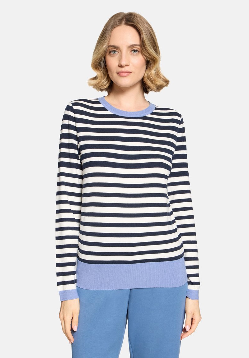 Femme aux cheveux blonds courts portant un pull rayé noir et blanc avec des poignets et un ourlet bleu clair, assorti à un pantalon bleu clair.