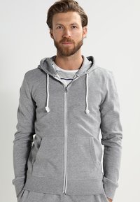 Grå zip-up hoodie i mjukt tyg med dragskohuva, framfickor och en avslappnad passform. Har vita dragskoaccenter.