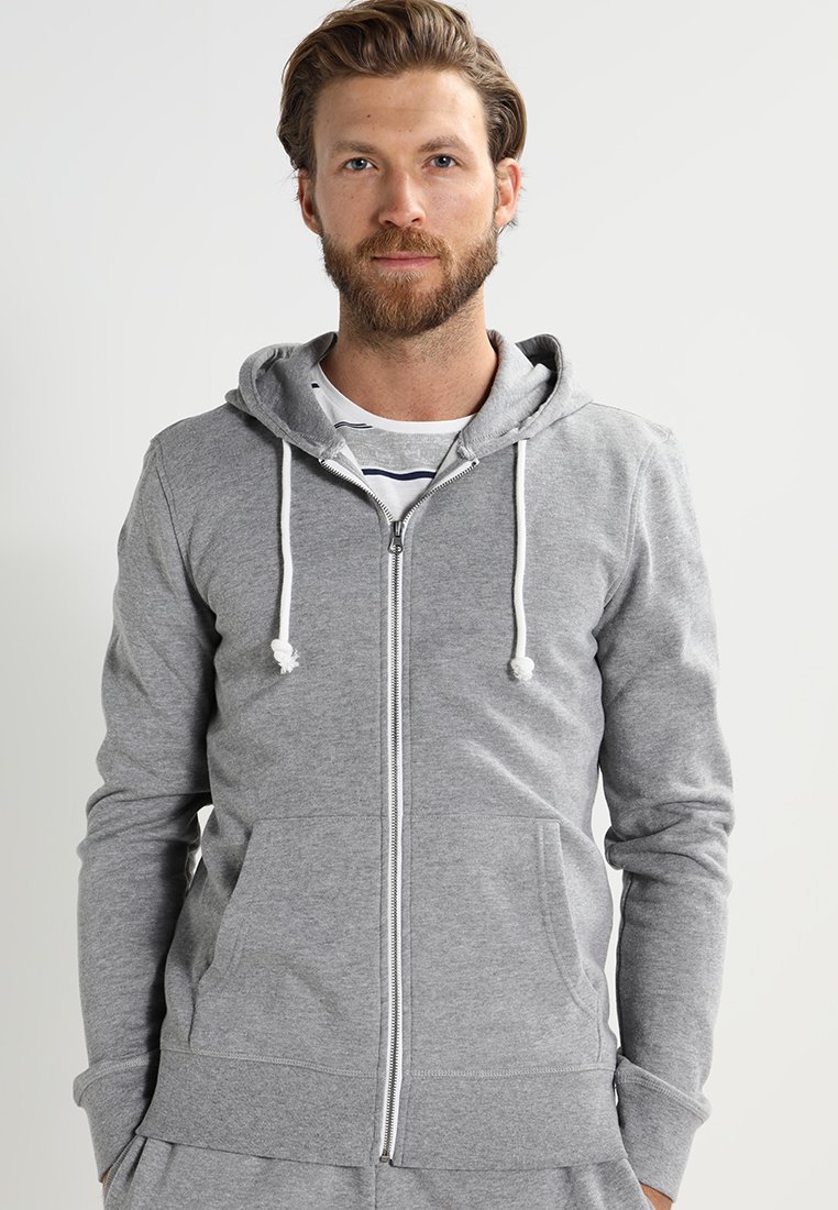 Grå zip-up hoodie i mjukt tyg med dragskohuva, framfickor och en avslappnad passform. Har vita dragskoaccenter.
