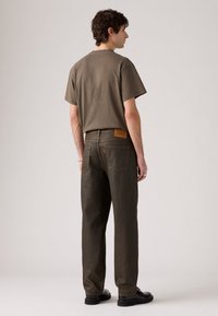 Moški obrnjeni stran, oblečen v rjavo majico, temno rjave kavbojke, črne čevlje, srebrno zapestnico in prstan, na enobarvni svetli podlagi.