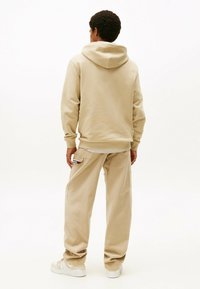 Beige Kapuzenpullover mit Kordelzug, kombiniert mit lockeren beigen Hosen mit einer Gesäßtasche. Strukturiertes Material, minimalistische Details.