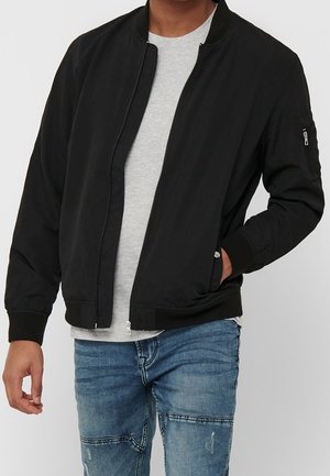 Un modèle cadré partiellement porte un blouson bomber noir zippé sur un T-shirt gris et un jean bleu, une main dans une poche.