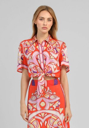 Femme portant un crop top boutonné à manches courtes avec un motif rouge vif, rose et violet, noué à la taille, et une jupe taille haute assortie.