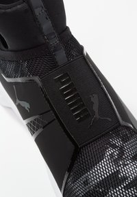 Svart sportskor med ett meshin övre, glansiga detaljer och texturerade mönster med en framträdande logotyp på sidan. Vit sula som kontrasterar.