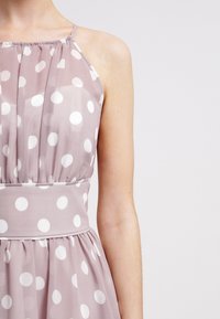Světlán růžové šaty s velkými bílými puntíky, s halter výstřihem, plisovaným živůtkem a přiléhavým pasovým dílem. Průsvitný materiál s měkkou texturou.