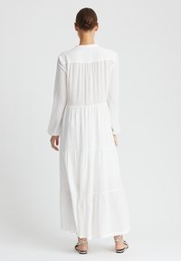 Robe maxi blanche à manches longues avec un design à niveaux, en tissu texturé et à col rond. Elle présente une coupe décontractée et un ourlet volantant.