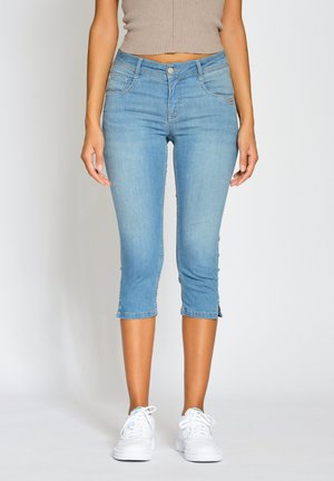 Femme portant un jean skinny court bleu clair, un top beige sans manches et des baskets blanches, debout devant un fond uni.