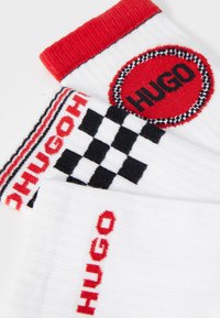 Calze di cotone bianche con accenti rossi, un logo circolare con la scritta "HUGO" e un motivo a scacchiera sul golf.