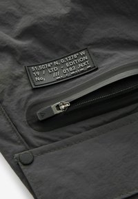 Tissu en nylon noir avec une poche zippée et une étiquette texturée avec des coordonnées et des détails d'édition. Les coutures le long des bords sont visibles.