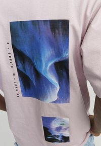 Rosa bomulls-t-shirt med två blå och lila abstrakta grafiska tryck på baksidan, med geografiska koordinater tryckta bredvid.