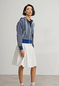 Modrá mikina na zips s vzorom a bielymi akcentmi, kombinovaná s bielou plisovanou sukňou. Modelka stojí v neutrálnom prostredí a predvádza outfit.