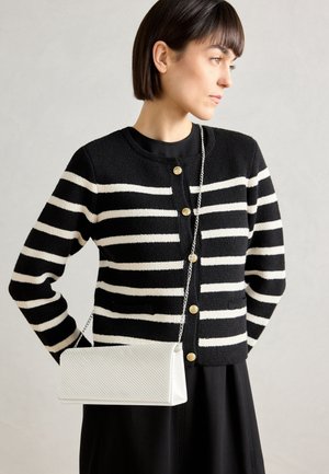 Mujer con cárdigan de rayas horizontales en blanco y negro con botones dorados, vestido negro y bolso blanco texturizado con asa de cadena plateada.