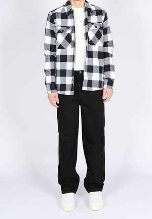 Homme portant une chemise à carreaux noir et blanc, un t-shirt blanc, un pantalon noir et des baskets blanches se tenant devant un arrière-plan uni.