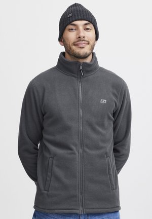 Gray fleece jacket with a high collar, full zip, and two front pockets. The model wears a matching dark knit beanie.