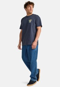 Camiseta de algodón azul marino para hombre, de manga corta y con gráfico en el frente, combinada con unos pantalones vaqueros de denim azul holgados y zapatillas negras.