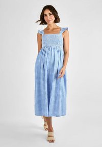 Vestido maxi a cuadros en azul y blanco con un cuerpo fruncido, tirantes con volantes y una falda fluida. Se lleva con sandalias de tacón de color claro.