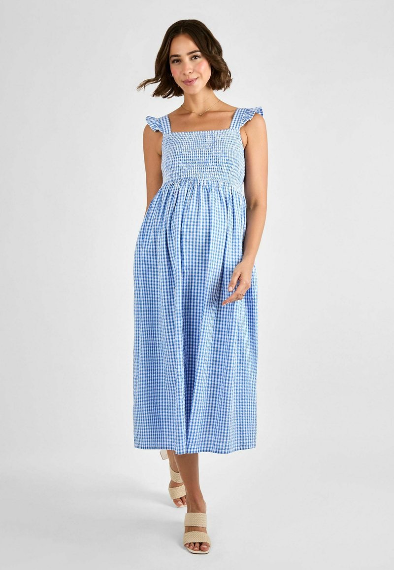 Vestido maxi a cuadros en azul y blanco con un cuerpo fruncido, tirantes con volantes y una falda fluida. Se lleva con sandalias de tacón de color claro.