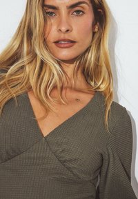 Žalios alyvuogės spalvos, tekstūruota viršutinė dalis su ilgomis rankovėmis, turinti žemą V formos iškirptę ir širdies tatuiruotę ant krūtinės. Lygus odos paviršius ir laisvi plaukai.