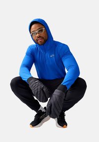 Blaues langärmliges Sportoberteil mit Kapuze, kombiniert mit schwarzen Hosen und gepolsterten grauen Handschuhen, getragen mit schwarzen Sneakers mit orangenen Akzenten.