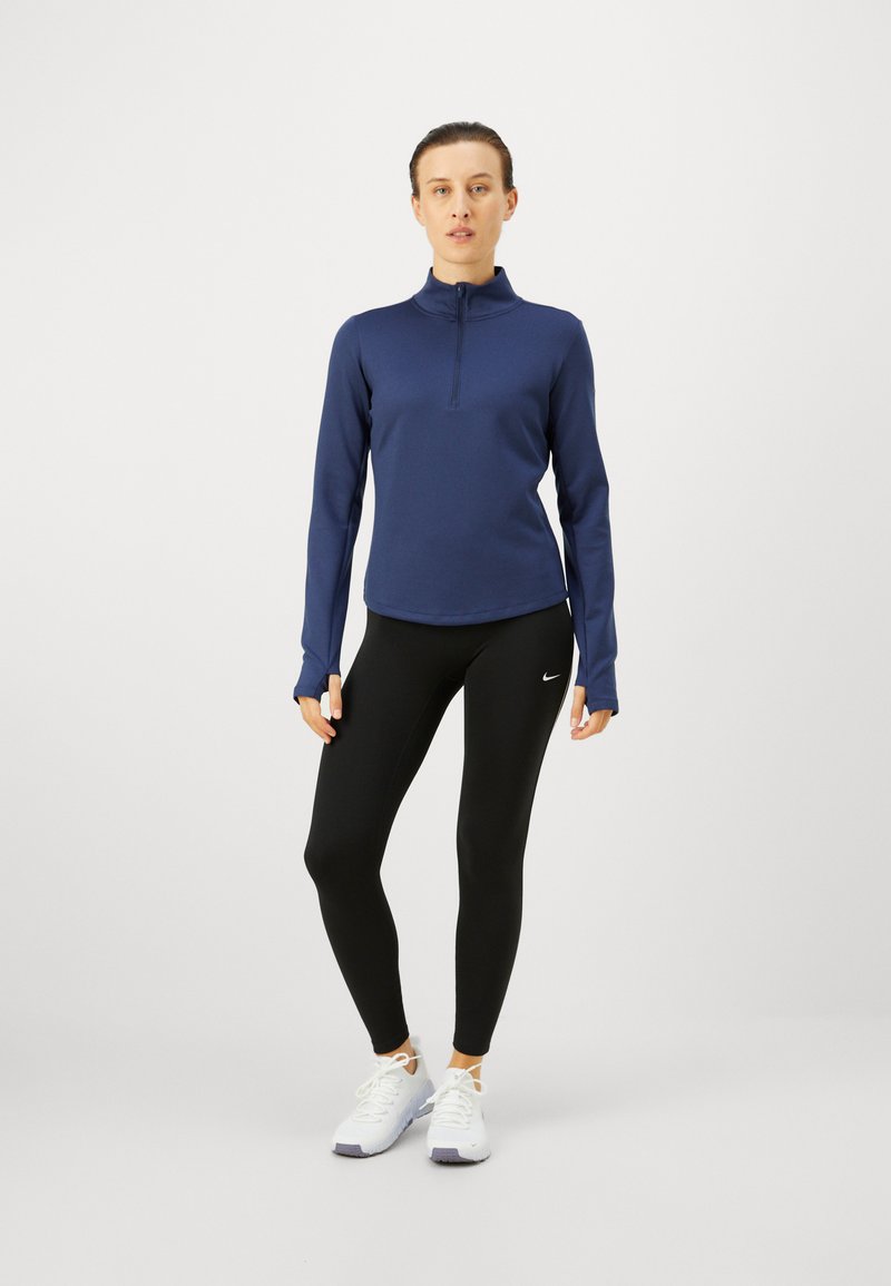 Marineblå langærmet pullover med høj krave og kvart lynlås, parret med sorte tights og hvide sneakers, der har en diskret logo-detalje.