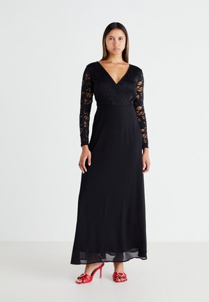 Black long-sleeve dress with a lace bodice, V-neckline, and flowing skirt. Paired with red heeled sandals featuring decorative accents.