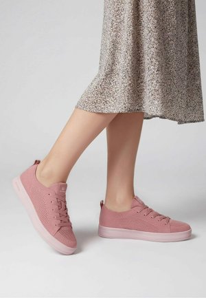 Benen met een beige bloemenrok en roze gebreide sneakers met veters, staand op een effen lichte achtergrond.