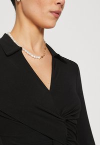 Svart omslagsblus med v-ringning och en samlad detalj. Ackompanjerad av ett halsband med pärlor och guldkedja, mjuk textur.