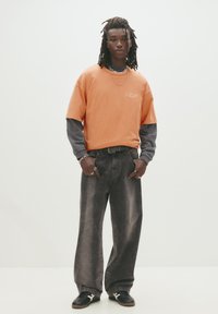 Orange oversized t-shirt lagd over en mørk langærmet skjorte, parret med falmede sorte brede jeans og sorte sko. Sølv smykker som accent.