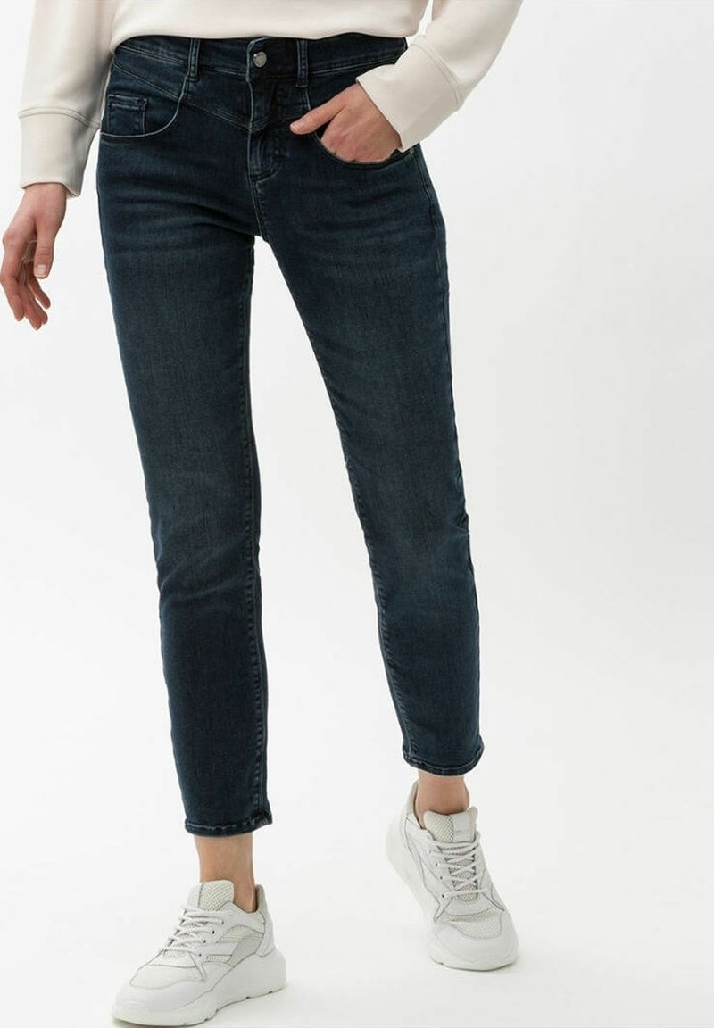 Personne portant un jean skinny bleu foncé et des baskets blanches, avec une main dans la poche, debout devant un fond uni.