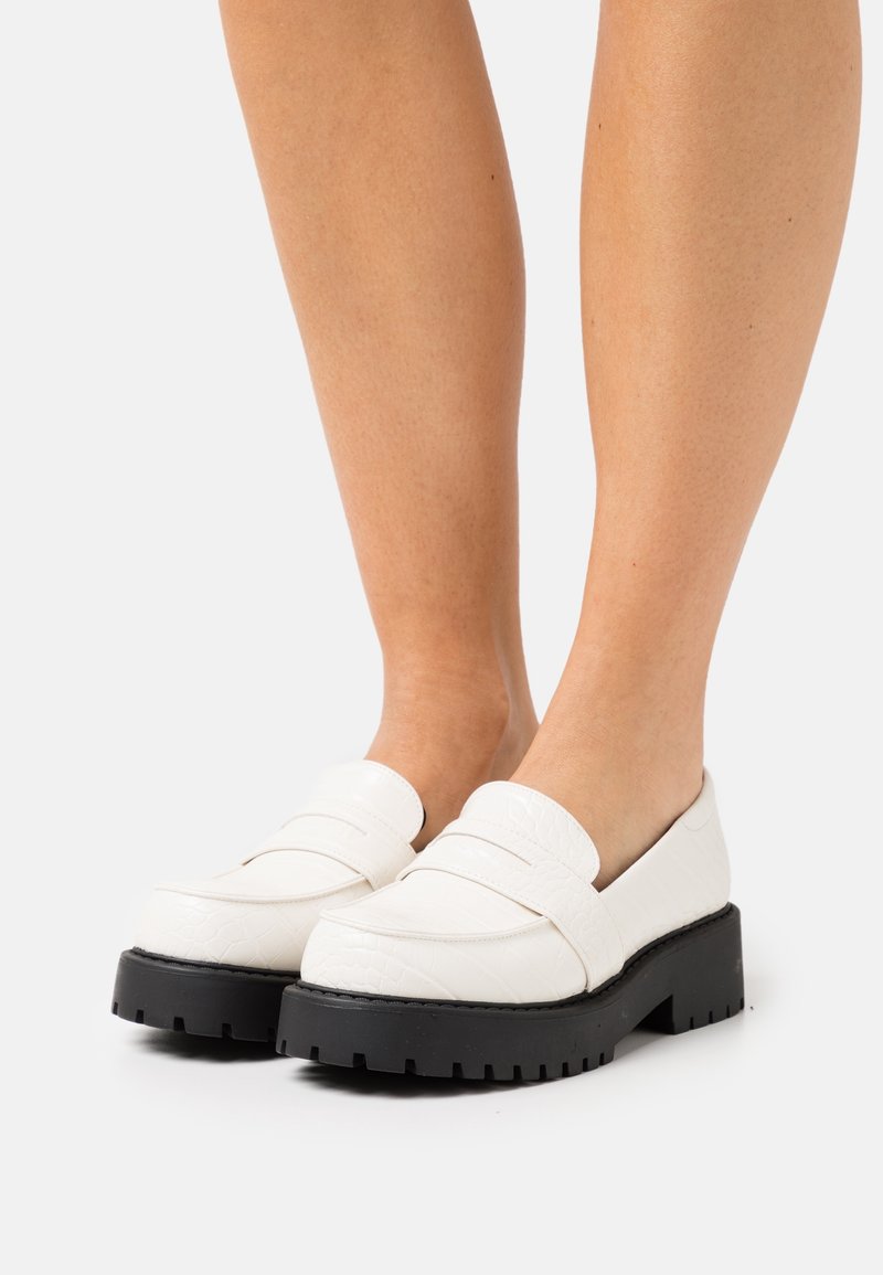 Femme portant des mocassins texturés blancs avec des semelles épaisses à crampons noires, debout sur un fond gris clair uni.