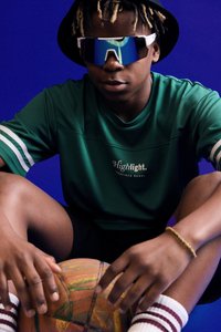 Grünes Sportshirt mit weißen Streifen, schwarze Shorts, reflektierende Sonnenbrille und einen bunten Basketball, vor einem blauen Hintergrund.