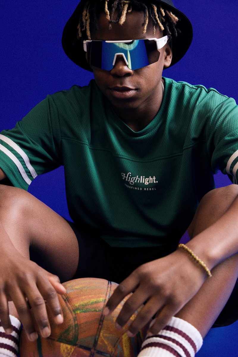 Grünes Sportshirt mit weißen Streifen, schwarze Shorts, reflektierende Sonnenbrille und einen bunten Basketball, vor einem blauen Hintergrund.