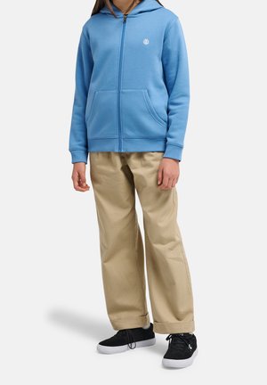 Osoba ubrana w jasnoniebieską bluzę na zamek, beżowe szerokie spodnie oraz czarne trampki z białymi podeszwami, stojąca na białym tle.