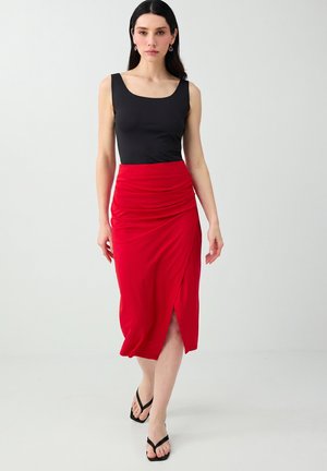 Donna che cammina in avanti indossando un top nero senza maniche, una gonna midi rossa con spacco laterale e sandali infradito neri su sfondo bianco.
