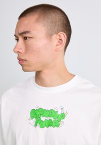 Ung mann med kort hår iført en hvit skjorte med grønn "RIPNDIP PUMA" tekst og tegneserieaktige hvite katter med blomster.