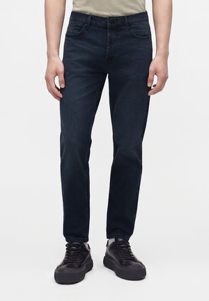 Homme portant un jean slim bleu foncé et des baskets en cuir noir, debout devant un fond clair uni.