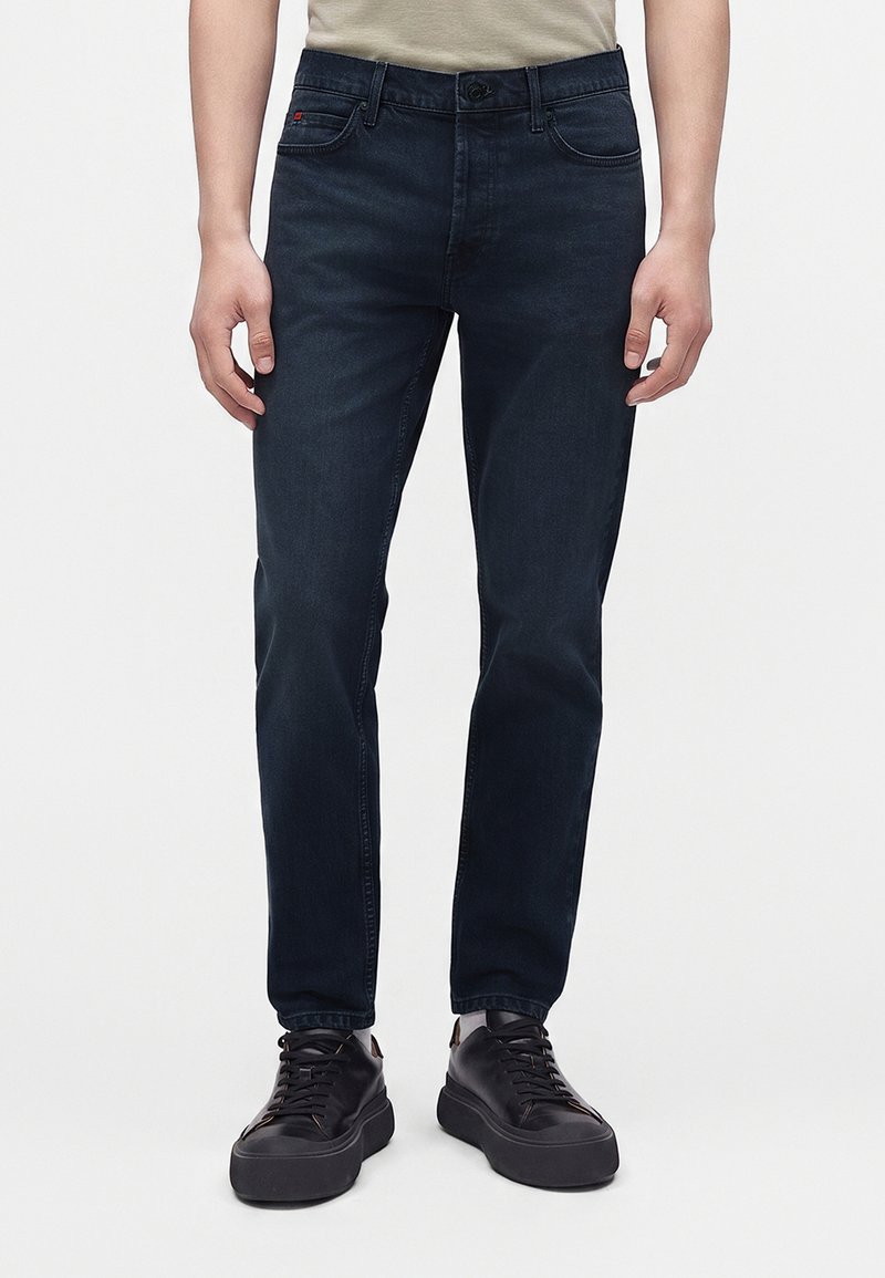Homme portant un jean slim bleu foncé et des baskets en cuir noir, debout devant un fond clair uni.