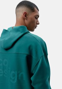 Tyrkysová mikina s uvoľneným strihom, s kapucňou, s embosovaným textom a kontrastným červeným akcentom na zadnom golieri. Mäkko zmes bavlny.