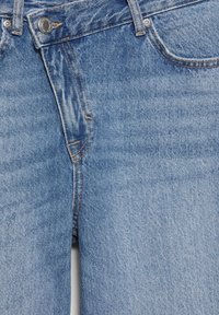 Lyseblå denimjeans i nærbillede, der viser lynlåsgylp, metal knap, forreste lomme og bæltestropper med synlige syningsdetaljer.