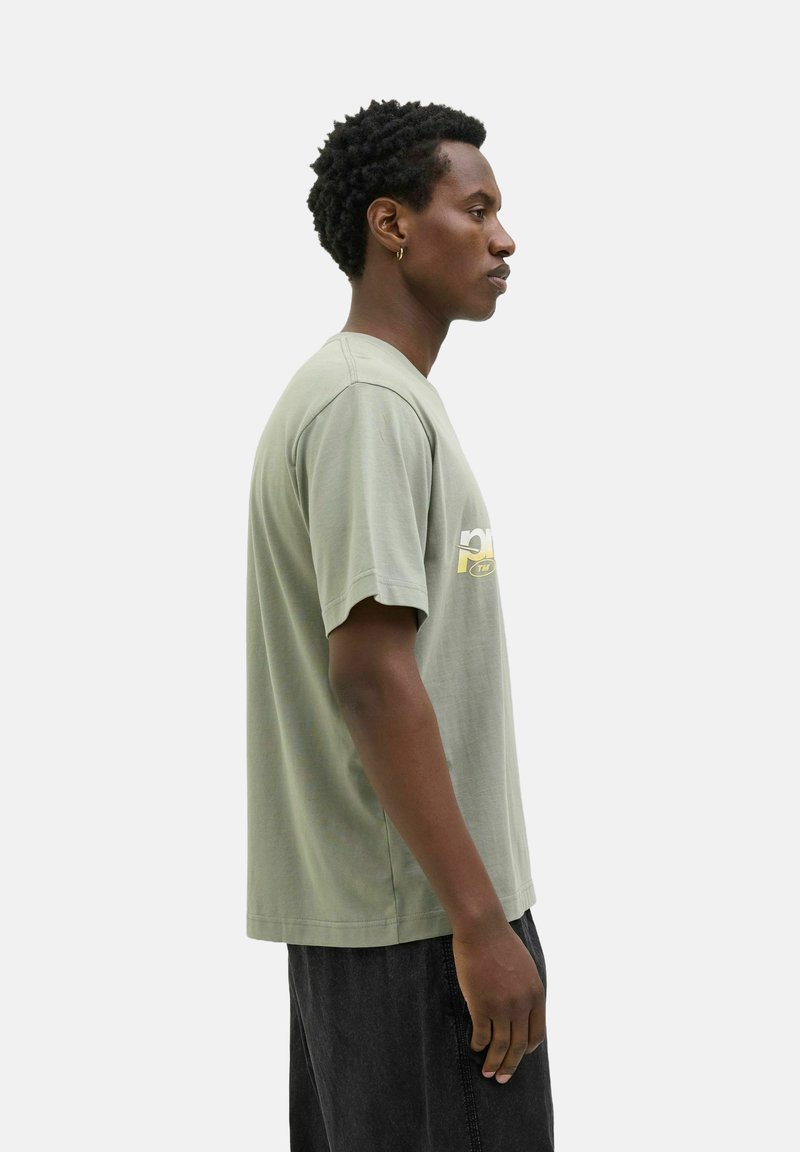 Grünes Baumwoll-T-Shirt mit kurzen Ärmeln, rundem Ausschnitt und einem gelben Grafikdruck auf der Brust. Getragen mit schwarzen Hosen.