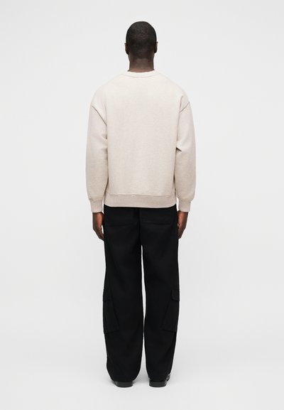 Άνδρας που στέκεται με την πλάτη, φοράει μπεζ φούτερ και μαύρο παντελόνι εργασίας, μπροστά σε απλό φωτεινό φόντο.