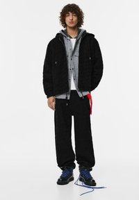 Černá texturovaná zipová bunda přes šedou kostkovanou košili a bílém tričku, doplněná černými kalhotami a černými botami s modrými tkaničkami.