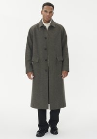 Uomo che indossa un lungo cappotto a quadri marrone, un dolcevita bianco, pantaloni neri e scarpe nere, in piedi contro uno sfondo bianco uniforme.