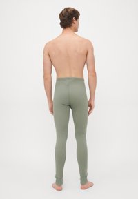 Uomo in piedi a piedi nudi, girato di spalle, che indossa pantaloni attillati di colore verde chiaro in un contesto con sfondo bianco semplice.