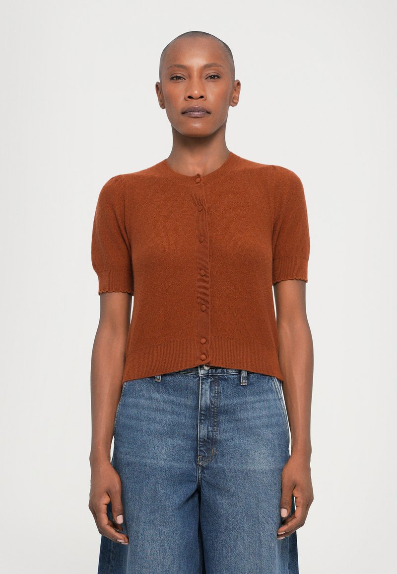 Brown short-sleeve cardigan with round neckline, button front, and ribbed hem. Worn over high-waisted blue jeans with a relaxed fit.