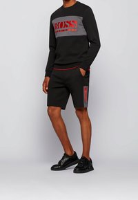 Felpa nera con pannello grigio e logo rosso, abbinata a pantaloncini neri con accenti rossi e grigi. Tessuto strutturato e vestibilità casual.