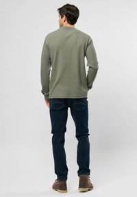 Olijfgroene katoenen top met lange mouwen en een textuurpatroon, gecombineerd met donkerblauwe jeans en bruine schoenen. Achteraanzicht, effen achtergrond.